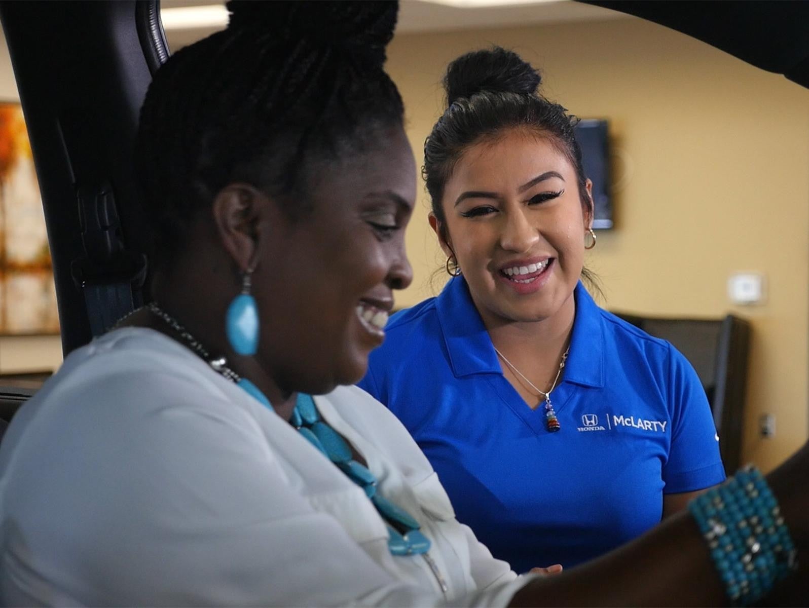 An employee showcases a car to a customer, who explores its features and comfort by sitting inside.